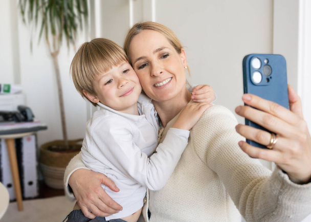 Sonriente joven madre e hijo pequeño usando el teléfono juntos, abrazando, en casa, madre feliz con adorable niño tomando selfie, disfrutando del tiempo libre con gadget - Foto, Imagen
