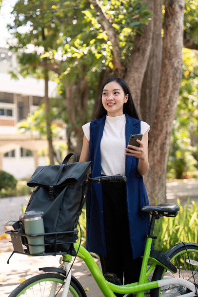 Mujer de negocios asiática con bicicleta usando smartphone y fuera del edificio de oficinas. Las mujeres que viajan en bicicleta van a trabajar. Vehículo ecológico, concepto de estilo de vida sostenible. - Foto, Imagen