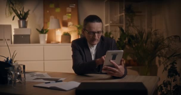 Starszy menedżer omawiający rozmowy wideo na tablecie cyfrowym podczas pracy do późna w miejscu pracy - Materiał filmowy, wideo