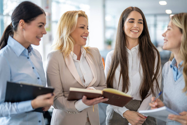 Multi-aged business women with paper documents standing in office interacting with each other, brainstorming creating sales strategy for their company. - Photo, Image