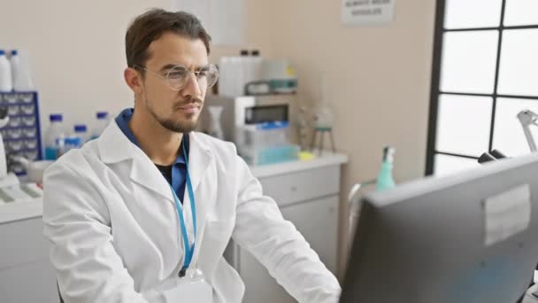 Um jovem cientista hispânico com barba trabalha atentamente em um computador em um ambiente de laboratório, exalando profissionalismo. - Filmagem, Vídeo