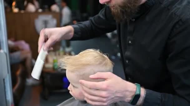 A férfi fodrász fésülködéssel és ollóval vágja a kisfiú haját. Egy felnőtt férfi vágja le a gyerekét, valószínűleg borbély. Professzionális fodrász és aranyos ügyfél a modern fodrászatban. - Felvétel, videó
