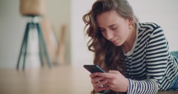 Felice giovane donna sms attraverso il telefono cellulare mentre si trova sul pavimento in soggiorno a casa - Filmati, video