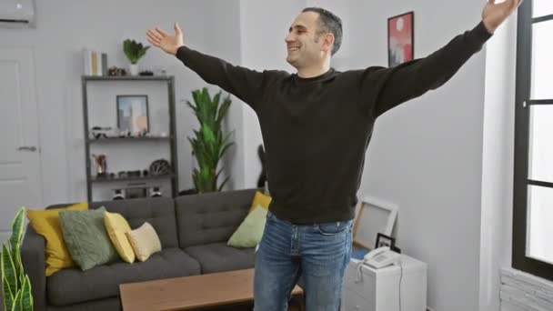 Ευτυχισμένος άντρας που απλώνει τα χέρια του σε εσωτερικούς χώρους κατά τη διάρκεια μιας χαλαρής ημέρας στο μοντέρνο διαμέρισμά του, μεταφέροντας ένα χαλαρό τρόπο ζωής. - Πλάνα, βίντεο