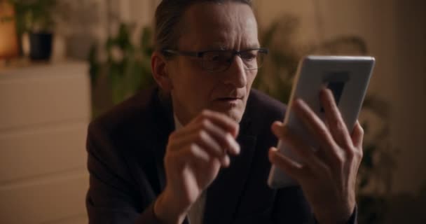 Vakava vanhempi liikemies silmälaseissa lukemassa digitaalista tablettia työskennellessään myöhään illalla toimistossa - Materiaali, video