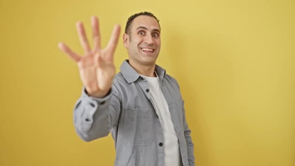 Alegre joven hispano con camisa muestra un cartel de "cuatro" manos, apuntando hacia arriba con confianza, aislado sobre un fondo amarillo feliz - Imágenes, Vídeo