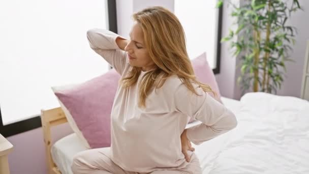 Una joven dolorida en pijama agarrando su espalda baja mientras estaba sentada en una cama en el interior, representando malestar o dolor de espalda. - Metraje, vídeo
