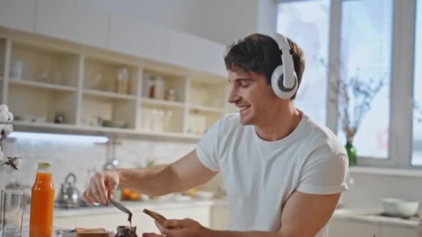 現代のキッチンのクローズアップで好きな歌に沿ってヘッドフォンで踊る料理人. 屋内で音楽を聴くイヤホンで朝食を準備する楽しい男. 自宅でトーストを作るおかしいメラノーマニア. - 映像、動画