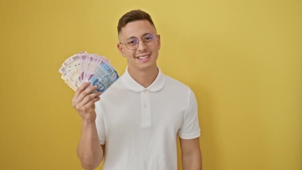 Wesoły młody, latynoski mężczyzna z radością wskazujący kciukiem na wiązkę meksykańskich banknotów peso, uśmiechnięty szeroko z otwartymi ustami na odizolowanym żółtym tle. - Materiał filmowy, wideo