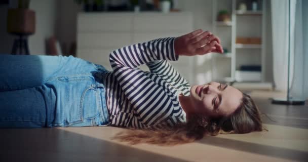 Uśmiechnięta młoda kobieta w koszulce w paski leżąca na podłodze pod słońcem w domu - Materiał filmowy, wideo