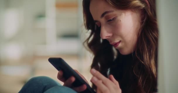 Młoda brunetka surfing internet na smartfonie do smsowania w salonie w domu - Materiał filmowy, wideo