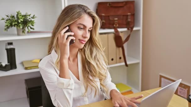 Beautiful, confident and smiling, a young blonde businesswoman masterfully manages work talks on her laptop and smartphone, making success look elegant, all from the comfort of her indoor office. - Footage, Video