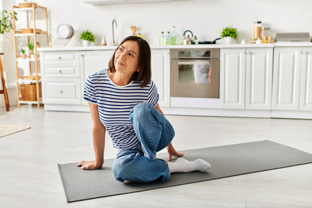 Uma mulher bonita madura em homewear aconchegante praticando ioga em um tapete em uma cozinha iluminada pelo sol. - Foto, Imagem