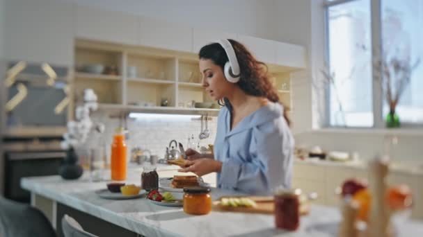Entspannte Frau kocht in Kopfhörer stehend gemütliche Küche. Attraktive junge Mädchen hören Musik im Headset und verteilen zu Hause Schokoladenpaste auf Toast. Sorglose Dame bereitet Mittagessen zu und genießt Gesang. - Filmmaterial, Video
