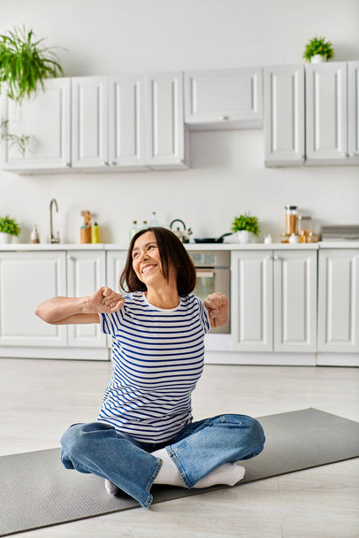 Egy érett nő kényelmes otthoni viseletben jógázik egy szőnyegen a konyhájában.. - Fotó, kép