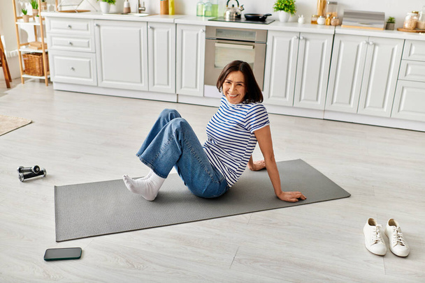 Reife Frau in Hausbekleidung praktiziert Yoga auf einer Matte in einer gemütlichen Küche. - Foto, Bild