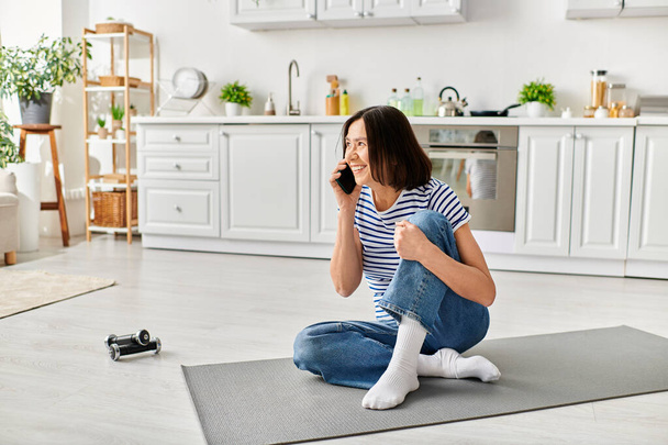 Una mujer madura en ropa de casa acogedora sentada en el suelo, comprometida en una conversación telefónica. - Foto, Imagen