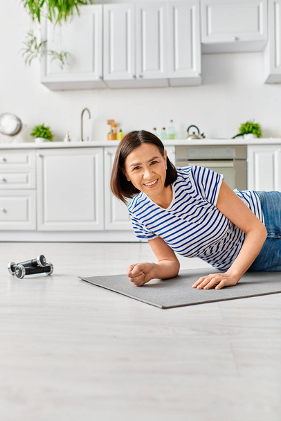 Une femme mature en tenue de maison pratique le yoga sur un tapis dans une cuisine ensoleillée. - Photo, image
