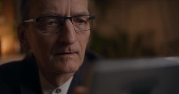 Fechamento do empresário sênior sério em óculos lendo no tablet digital enquanto trabalhava até tarde da noite no escritório - Filmagem, Vídeo