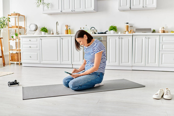 Reife Frau in gemütlicher Kleidung sitzt auf Yogamatte und benutzt Handy im ruhigen Morgenraum. - Foto, Bild