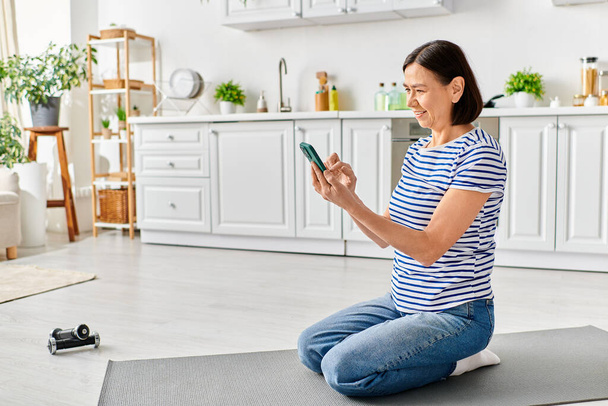 Reife Frau in gemütlicher Kleidung sitzt auf Yogamatte und konzentriert sich aufs Telefon. - Foto, Bild