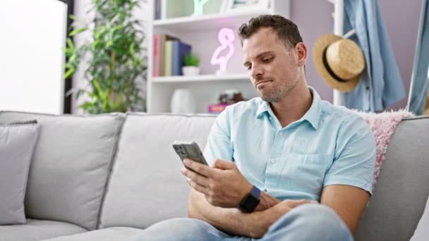 Bello uomo con barba utilizzando smartphone sul divano nel moderno soggiorno interno - Filmati, video