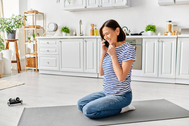 Evde oturan olgun bir kadın yoga minderinde otururken telefonuyla sohbet ediyor.. - Fotoğraf, Görsel