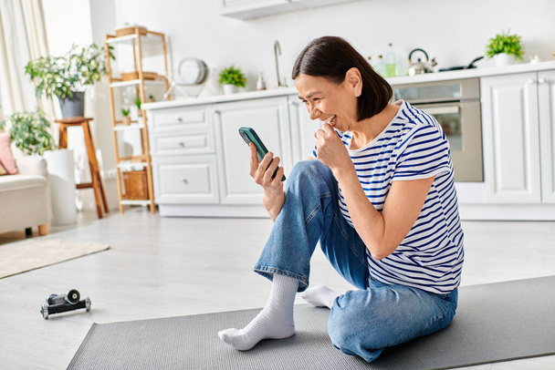 Una mujer madura y hermosa en ropa de casa acogedora se sienta en el suelo, absorta en su teléfono celular. - Foto, Imagen