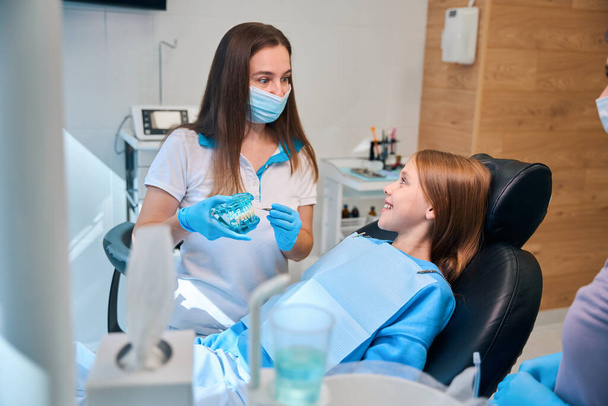 Mulher higienista se comunica durante uma consulta com um jovem paciente, ela tem um modelo da dentição em suas mãos - Foto, Imagem