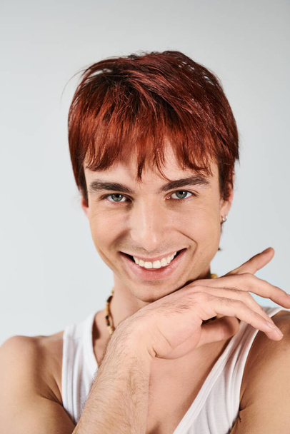 Een stijlvolle jongeman met rood haar die vol vertrouwen poseert in een wit topje tegen een grijze studio achtergrond. - Foto, afbeelding