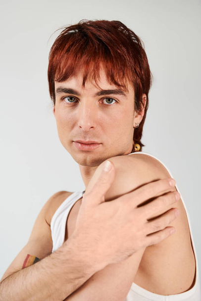 Un joven elegante con el pelo rojo posa con confianza en una camiseta blanca sobre un fondo gris de estudio. - Foto, imagen