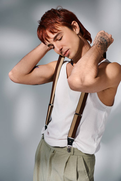 A fashionable young man with red hair poses confidently in a white shirt and suspenders against a grey studio backdrop. - Φωτογραφία, εικόνα