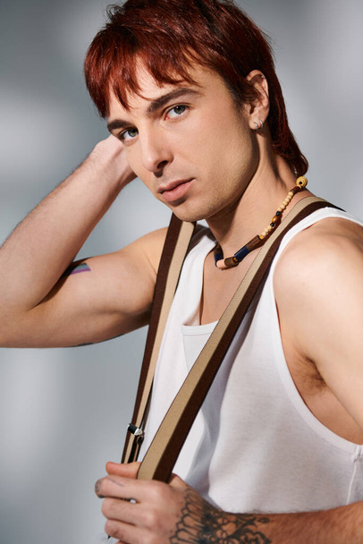 Egy elegáns, vörös hajú fiatalember pózol egy fehér trikóban, szürke stúdióháttérrel.. - Fotó, kép
