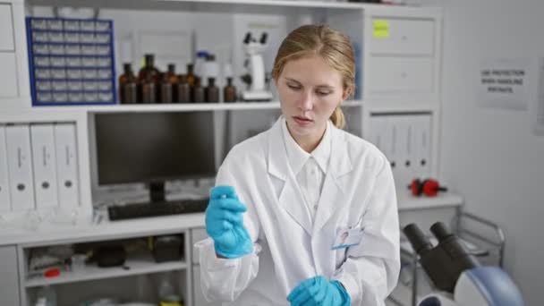 Een gefocuste vrouwelijke wetenschapper onderzoekt een reageerbuis in een laboratoriumomgeving, omringd door apparatuur en opbergkasten. - Video