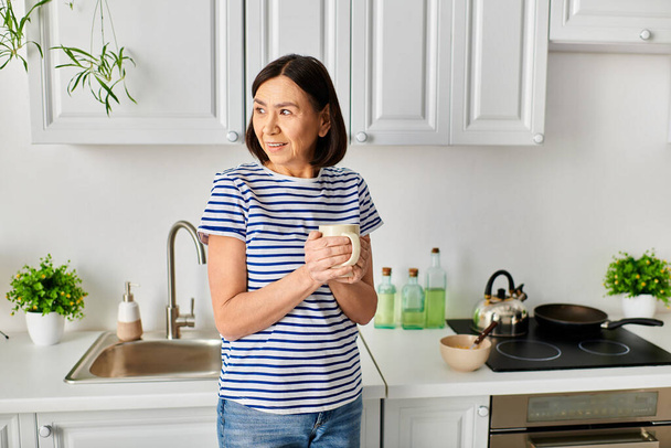 Eine reife Frau in gemütlicher Hauskleidung steht in einer Küche und hält eine Tasse. - Foto, Bild