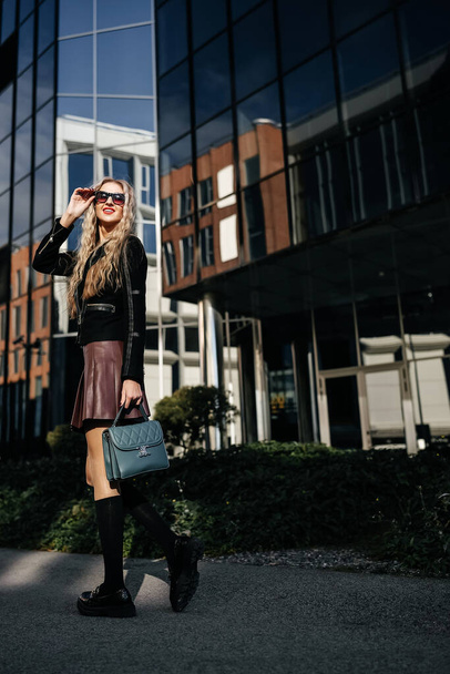 Fashionable young woman in black jacket wearing dark sunglasses posing on the city street. Blonde woman posing on glass building background. - Foto, immagini