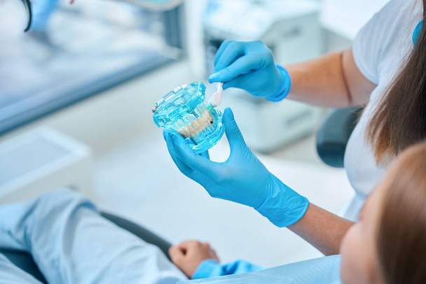 La doctora sostiene un modelo de dentición y un cepillo de dientes en sus manos, consulta a una adolescente en una clínica dental - Foto, Imagen