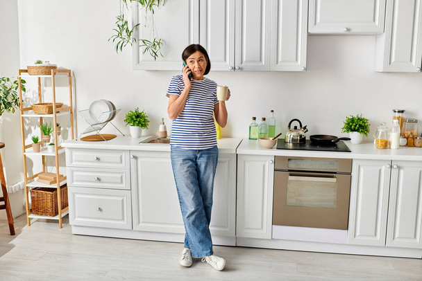 Una mujer en ropa de casa acogedora hablando en un teléfono celular en una cocina. - Foto, Imagen