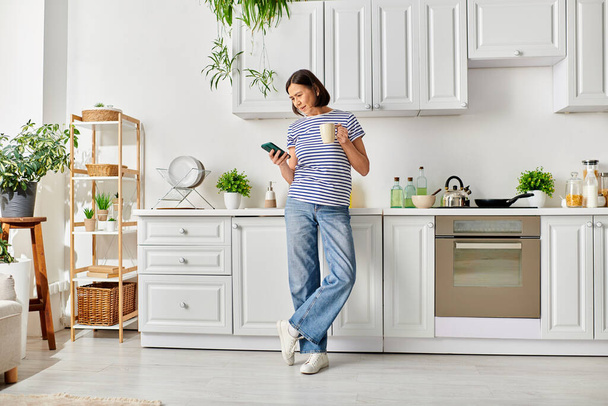 Uma mulher madura em roupa de casa, de pé em uma cozinha com um telefone celular. - Foto, Imagem