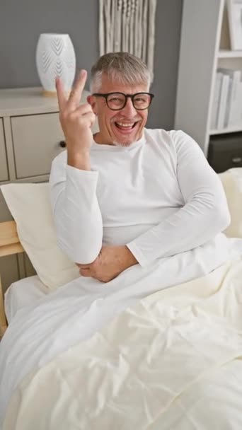 Fröhlicher grauhaariger Mann mittleren Alters, der im Schlafzimmer siegreich lächelt, selbstbewusst zwinkert und im Schlafanzug ein Zwei-Finger-Siegeszeichen blinkt - Filmmaterial, Video
