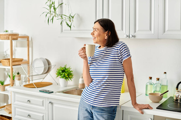 Зріла жінка насолоджується спокійним моментом на своїй кухні, тримаючи чашку кави. - Фото, зображення