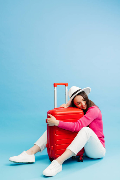 Happy young tourist woman sitting at the suitcase, hugging it and going to travel on holidays isolated on blue background - Photo, Image