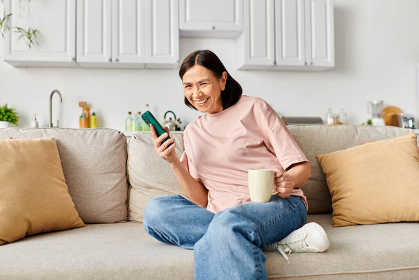 Зріла жінка в затишному домашньому вбранні насолоджується спокійним моментом на дивані з мобільним телефоном. - Фото, зображення
