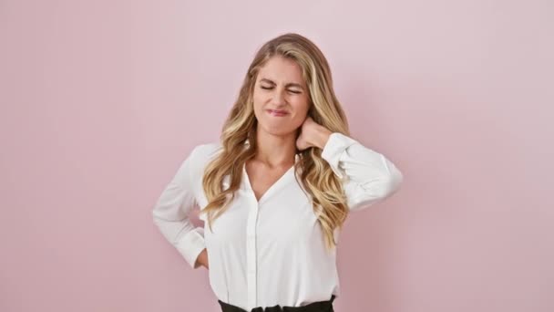 Pijnlijk gestresste jonge blonde vrouw met een shirt aan, die last heeft van nekpijn. staand masseert ze haar pijnlijke spieren over een roze, geïsoleerde achtergrond - Video