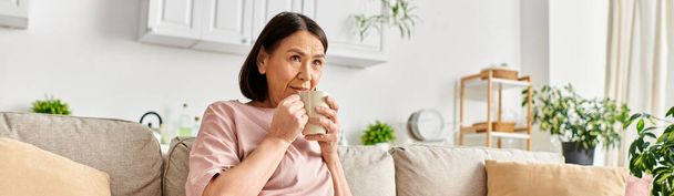 Μια ώριμη γυναίκα σε ζεστό homewear κάθεται σε έναν καναπέ, με χάρη πίνοντας από ένα φλιτζάνι. - Φωτογραφία, εικόνα