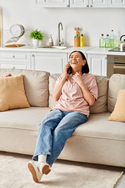 Reife Frau in Homewear spricht auf dem Handy, während sie auf der Couch sitzt. - Foto, Bild