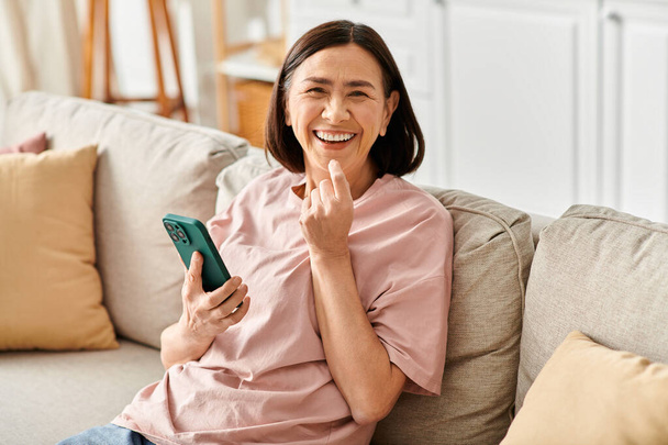 Μια ώριμη γυναίκα με ζεστά ρούχα κάθεται σε έναν καναπέ, κρατώντας ένα κινητό τηλέφωνο. - Φωτογραφία, εικόνα