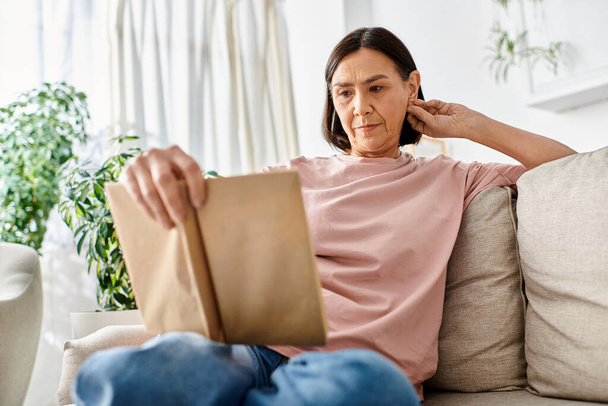 Eine reife Frau in bequemer Homewear sitzt auf einer Couch und ist in ein Buch vertieft. - Foto, Bild