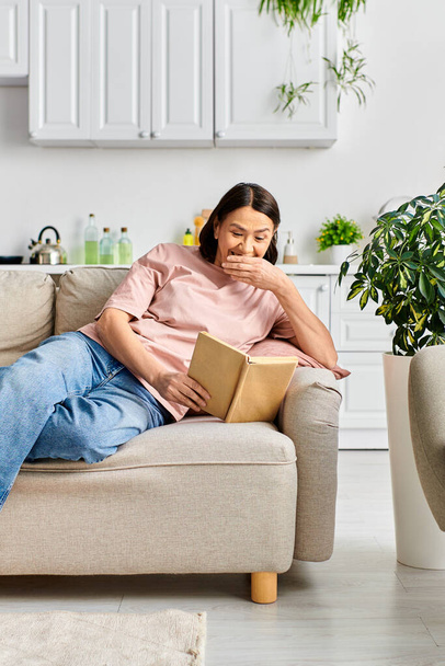 Žena v útulných domácích šatech sedící na gauči, hluboce zabraná do čtení knihy. - Fotografie, Obrázek