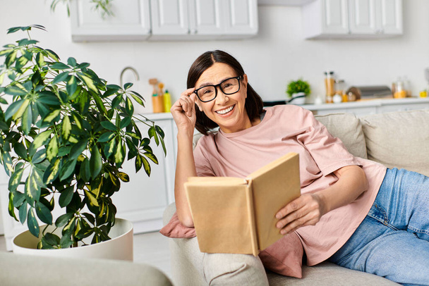 Eine reife Frau in kuscheliger Homewear entspannt sich auf einer Couch, eingetaucht in ein Buch. - Foto, Bild